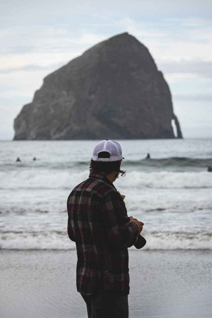 Garrett with camera in at hand looking out Kiwanda/Pacific Beach with a big rock in the distance.